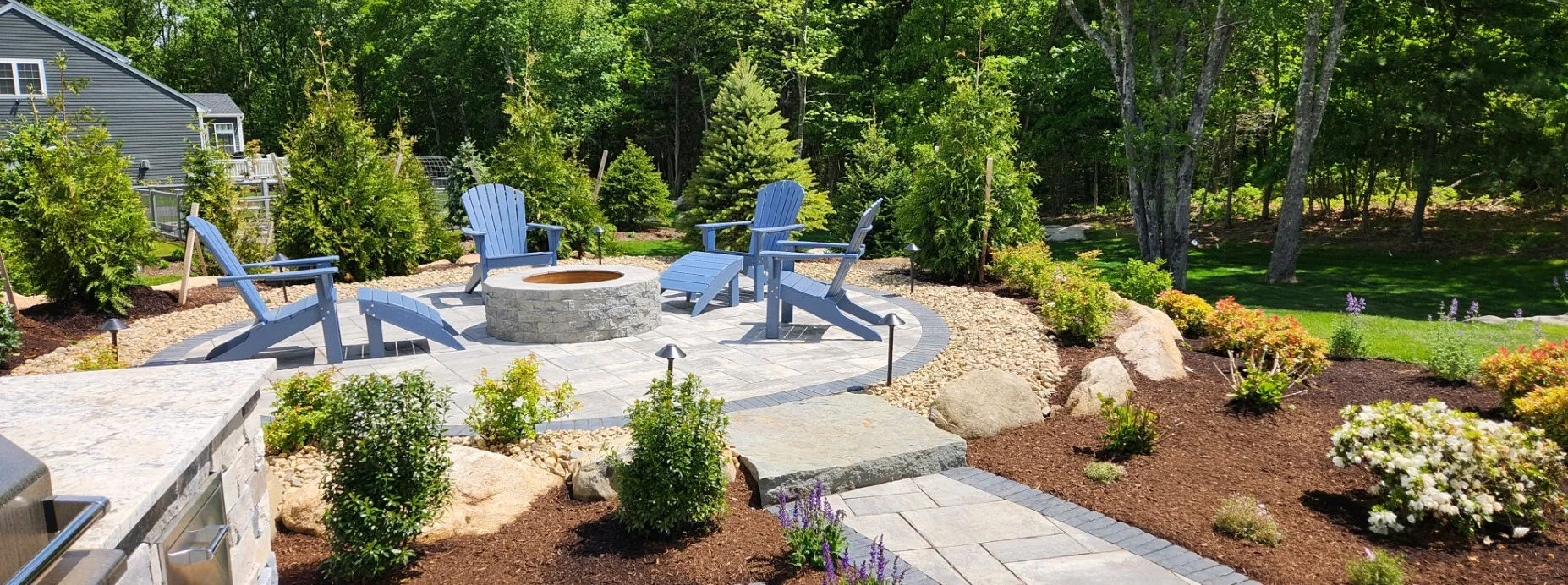 Backyard stone patio with round firepit, blue Adirondack chairs and fresh plantings
