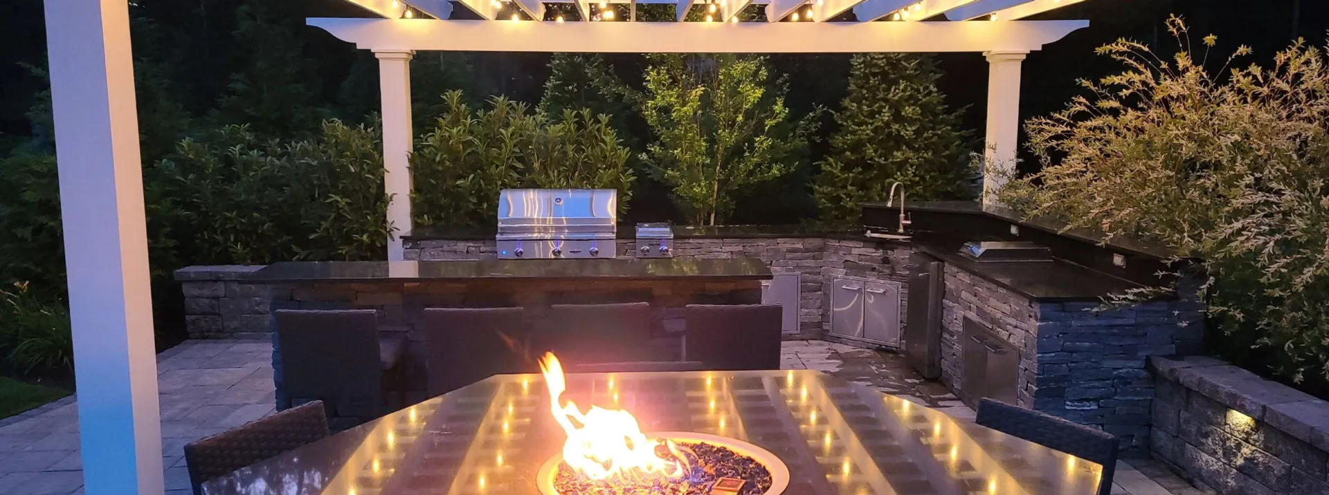 Firepit table and stone outdoor kitchen under a lit pergola